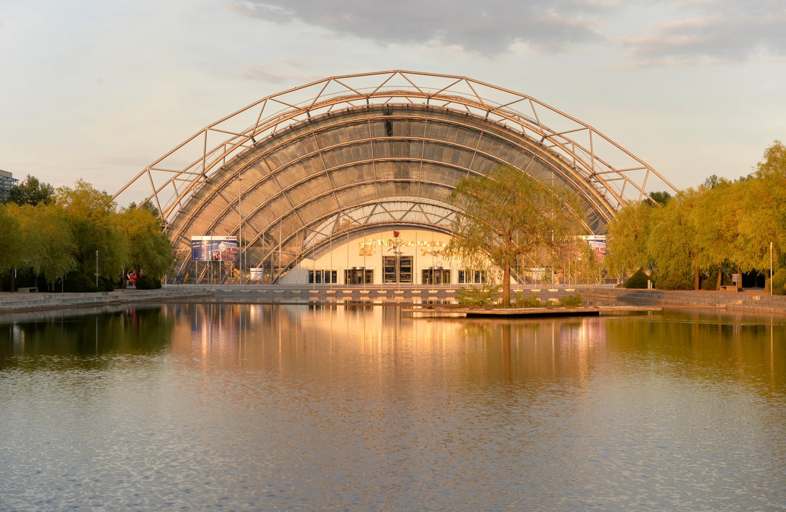 Die imposante Glas- und Stahlkuppel des Eingangsbereichs der Leipziger Messe spiegelt sich in einem ruhigen Teich bei goldenem Sonnenlicht. Umgeben von Bäumen, die das warme Licht reflektieren, bietet die Szene einen harmonischen Anblick.